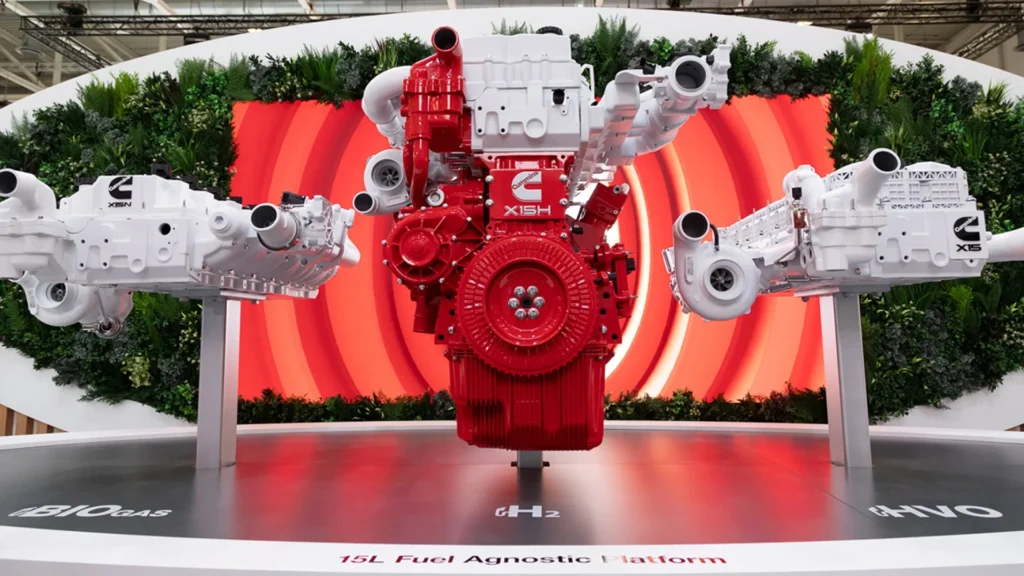 A red and white Cummins fuel-agnostic X-Series engine sits on a stage with a red background and greenery at an exposition